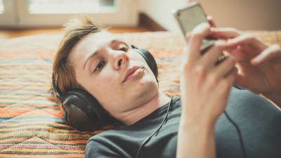 Kvinna med kort hår ligger på säng med hörlurar och lyssnar på nåt från mobilen.