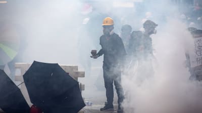 Demonstrant i Hongkong iförd gasmask och gul skyddshjälm står mitt i ett moln av tårgas.