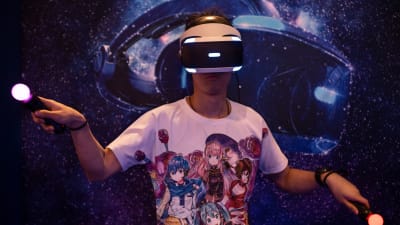 En man med VR-glas och kontroller i händerna ses spela ett spel. Han har på sig en t-shirt med anime-figurer.