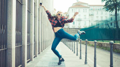 En kvinna som lyssnar på musik i hörlurar. Hon står på en gata och hoppar högt i luften. Ser glad ut. 