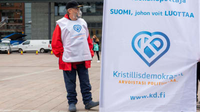 Kristdemokraternas kandidat på Narinkens torg bredvid en flagga med partiets logo.