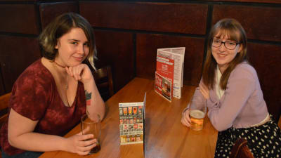 Två unga kvinnor sitter mittemot varandra vid ett bord i en pub.