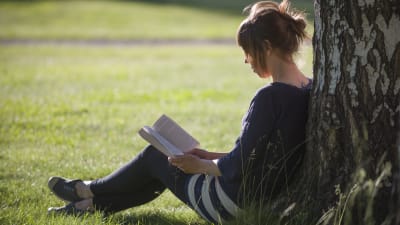 Nainen istuu nurmikolla, lukee kirjaa ja nojaa koivuun