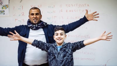 En vuxen man och en pojke står med utsträckta armar framför en skoltavla med finska ord
