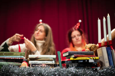 Två kvinnor sitter bakom högar av böcker och pysslar med presentpapper och -band. Kvinnorna är suddiga i bakgrunden, böckerna och julglitter i fokus.