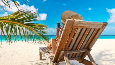 En kvinna i en solstol på en strand sitter och petar på en smarttelefon.