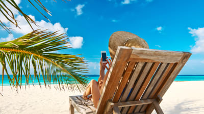En kvinna i en solstol på en strand sitter och petar på en smarttelefon.
