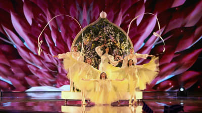 katerine duska uppträder med gulklädda balettdansare och en stor blomvägg bakom
