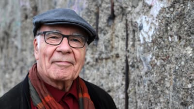 En äldre man med en skärmmössa på huvudet, glasögon, röd skjorta, svart kavaj och en färggrant rutig halsduk i orangenyanser tittar vänligt, men bestämt in i kameran. I bakgrunden är en grå mur.