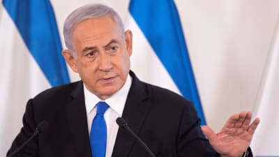 Netanjahu puhuu mikrofoniin. Hänellä on musta puku. valkoinen kauluspaita ja sininen kravatti. Taustalla näkyy Israelin sinivalkoinen lippu.
