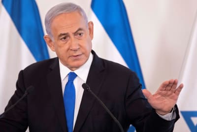 Netanjahu puhuu mikrofoniin. Hänellä on musta puku. valkoinen kauluspaita ja sininen kravatti. Taustalla näkyy Israelin sinivalkoinen lippu.