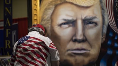 Man i fägrgglad skjorta med USA-flagga beundrar tavla av Donald Trump.