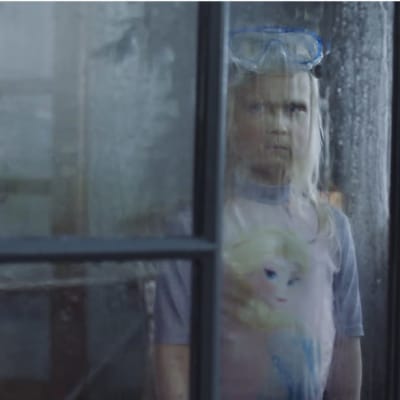 Vaaleahiuksinen tyttö katsoo ulos surullisena ikkunasta sateiseen säähän. Tytöllä on päällään isot uimalasit ja uimapuku.