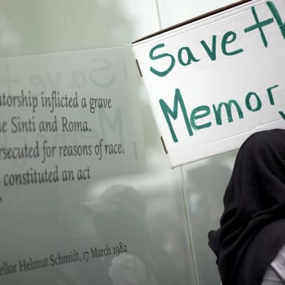 Mielenosoittaja pitelee pahvilaatikosta tehtyä kylttiä, jossa lukee englanniksi "pelastakaa muisto". Taustalla näkyy muistomerkin teksti, jossa liittokansleri Helmut Kohl tunnustaa romanien kansanmurhan. 