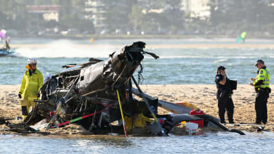 Pelastushenkilöt tutkivat törmäyksessä tuhoutunutta helikopteria rannalla.