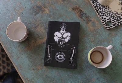 Kahden kahvikupin välissä on MSC Finland - Tom's Club -kirja, jonka kannessa on piirretty koppalakkinen ja nahkavaatteisiin pukeutunut lihaksikas mies.