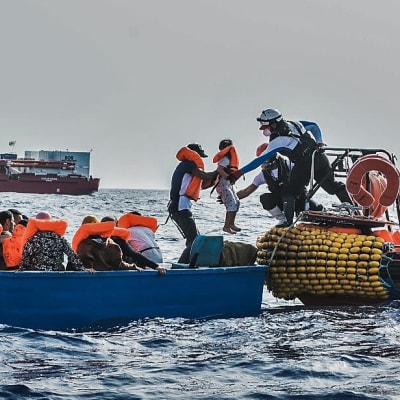 Hjälparbetare hjälper migranter ombord en orange gummibåt, fartyget Ocean Viking i bakgrunden. 