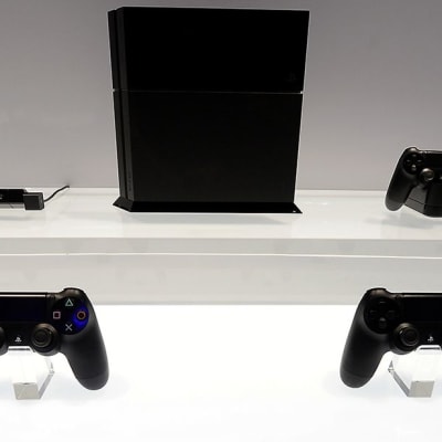 Väriltään musta playstation 4 -pelikonsoli ja  mustia peliohjaimia esillä E3-messuilla kirkkaan valkoista alustaa vasten.