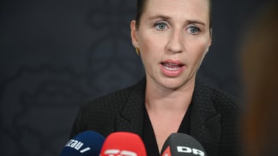Danmarks statsminister Mette Frederiksen i kostym talar inför pressen. I förgrunden syns tre mikrofoner på hennes bord. 