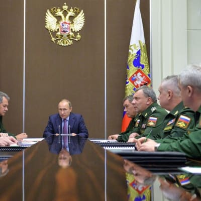 Vladimir Putin istuu pöydän päässä. Pöydän molemmin puolin istuu sotilaita.