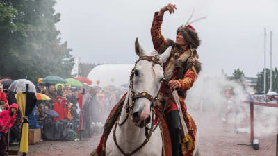 Keskiaikaiseen asuun pukeutunut nainen heilauttaa kättään valkoisen hevosen selässä. Taustalla savua ja yleisöä vesisateessa.