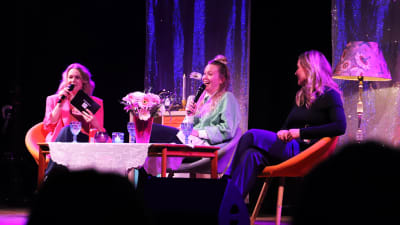 Tre kvinnor diskuterar på en scen framför publik.