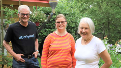 Tre personer, en man och två kvinnor, står på en grönskande gård i Raunistula.