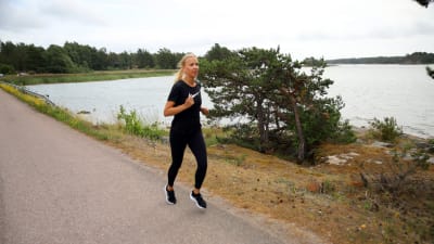 En kvinna med ljust hår i hästsvans, svart skjorta och svarta tights springer på en somrig asfaltväg med havet i bakgrunden.