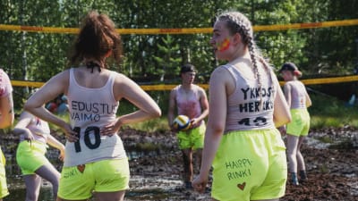 Unga kvinnor i gula shorts och vita linnen står till knäna insjunkna i en torvmosse och spelar kärrvolleyboll.