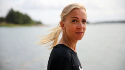 En kvinna med ljust hår i hästsvans och svart skjorta tittar rakt in i kameran. i bakgrundens syns havet och öar suddigt.