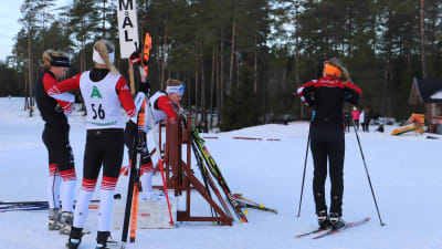 En grupp flickor på skidor står runt en stolpe där det står "mål".