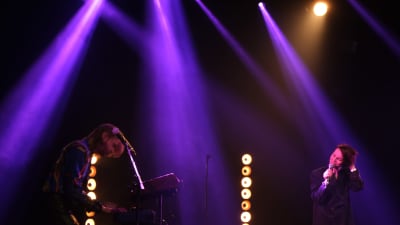 Man spelar keyboards, kvinna står med mikrofon på en scen. Starka ljus.