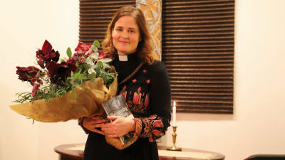 En kvinnlig präst står med en blombukett och en bok i famnen.