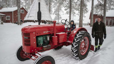 En liten pojke sitter på en röd gammal traktor, hans pappa tittar på.