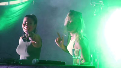 Två unga kvinnor står i ett dj-bås och dansar i ett grönt ljus. Den ena pekar rakt mot kameran.