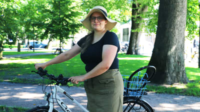 En kvinna med svart t-skjorta och hatt står och håller i en cykel.