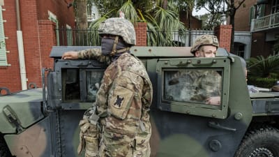 Soldater i kamouflagedräkt och munskydd står bredvid ett militärfordon.