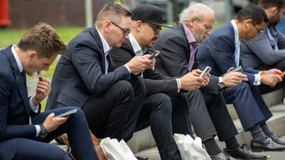 En rad män i kostym sitter på trottoarkanten och tittar på sina telefoner.