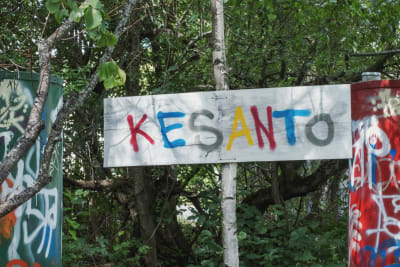 Skylt med texten "Kesanto" skriven med färggranna bokstäver. I bakgrunden lövträd.