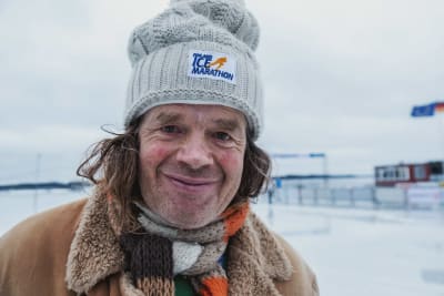 Äldre man klädd i vinterkappa, randig halsduk och mössa med texten "Finland Ice Marathon" i vintrigt landskap.