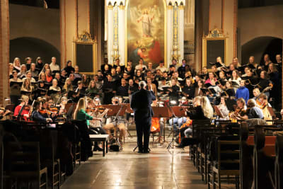 En man står med ryggen mot kameran och dirigerar en kör och en orkester i en kyrka. 