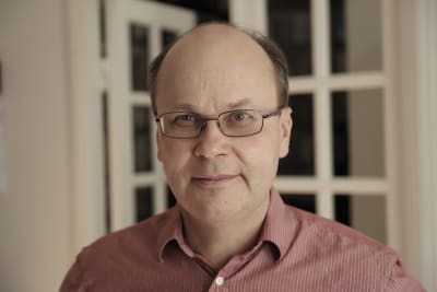 En man i röd skorta står i ett vitt rum. 