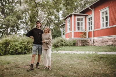 Två personer står bredvid varandra på en gräsmatta och ler. I bakgrunden syns ett rött hus.