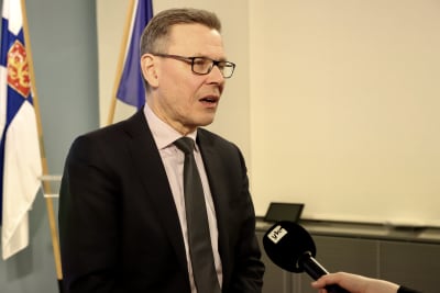 En man i kostym, slips och glasögon med Finlands statsflagga i bakgrunden intervjuas för TV. 