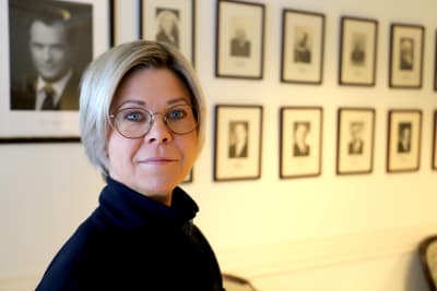 En kvinna med glasögon tittar in i kameran. I bakgrunden porträtt på Sauli Niinistö. 