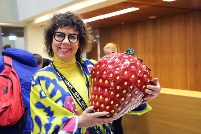 En kvinna med lockigt hår och färgglada kläder håller i en stor jordgubbe.