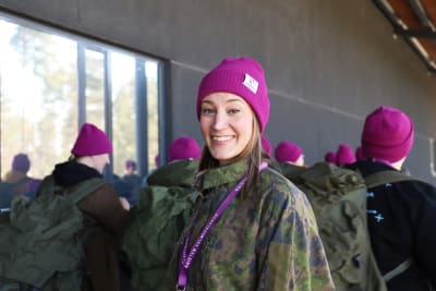 Kvinna med rosa mössa och militärjacka tittar rakt in i kameran och ler. 