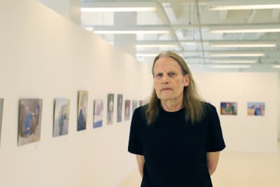 Kaj Stenvall står framför en vägg med en rad av tavlor. 