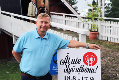 En man i turkos skjorta poserar vid en skylt med texten Ät ute.