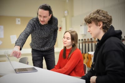 Musikläraren Tomi Norha pekar ut något på en laptop för gymnasieeleverna Oona Vahtera och Antti Lampi.